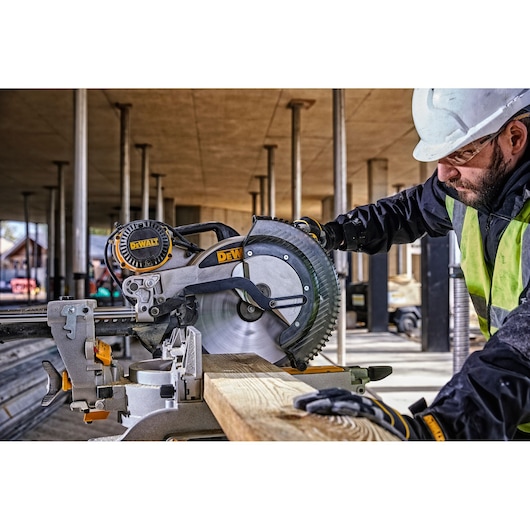 A DEWALT DWS780 miter saw being used to cut a wooden plank at a construction site. The operator is wearing a safety helmet, gloves, and a high-visibility vest. The background shows a partially constructed building with vertical pillars.