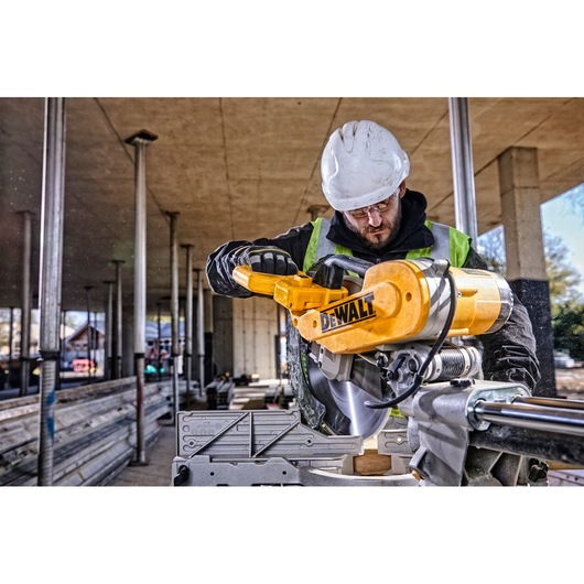 A person wearing a white safety helmet and gloves operates a yellow DEWALT DWS780 miter saw at a construction site. The background shows an unfinished building structure with columns and beams.