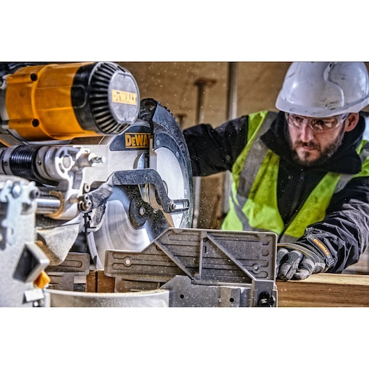 A DEWALT branded miter saw, model DWS780, is being used by a person wearing a safety vest, gloves, and a hard hat to cut a piece of wood at a construction site. The person's face is blurred for privacy.
