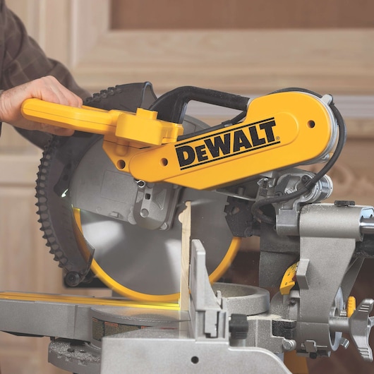 A person is using a DEWALT DWS780 miter saw to cut a piece of wood. The saw is yellow and gray, with the DEWALT logo prominently displayed. The wood is positioned on the cutting platform, and sawdust is visible.
