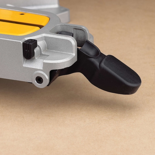 Close-up of a DEWALT miter saw component, showing a metallic part with a black plastic lever and a yellow detail, placed on a light brown surface.